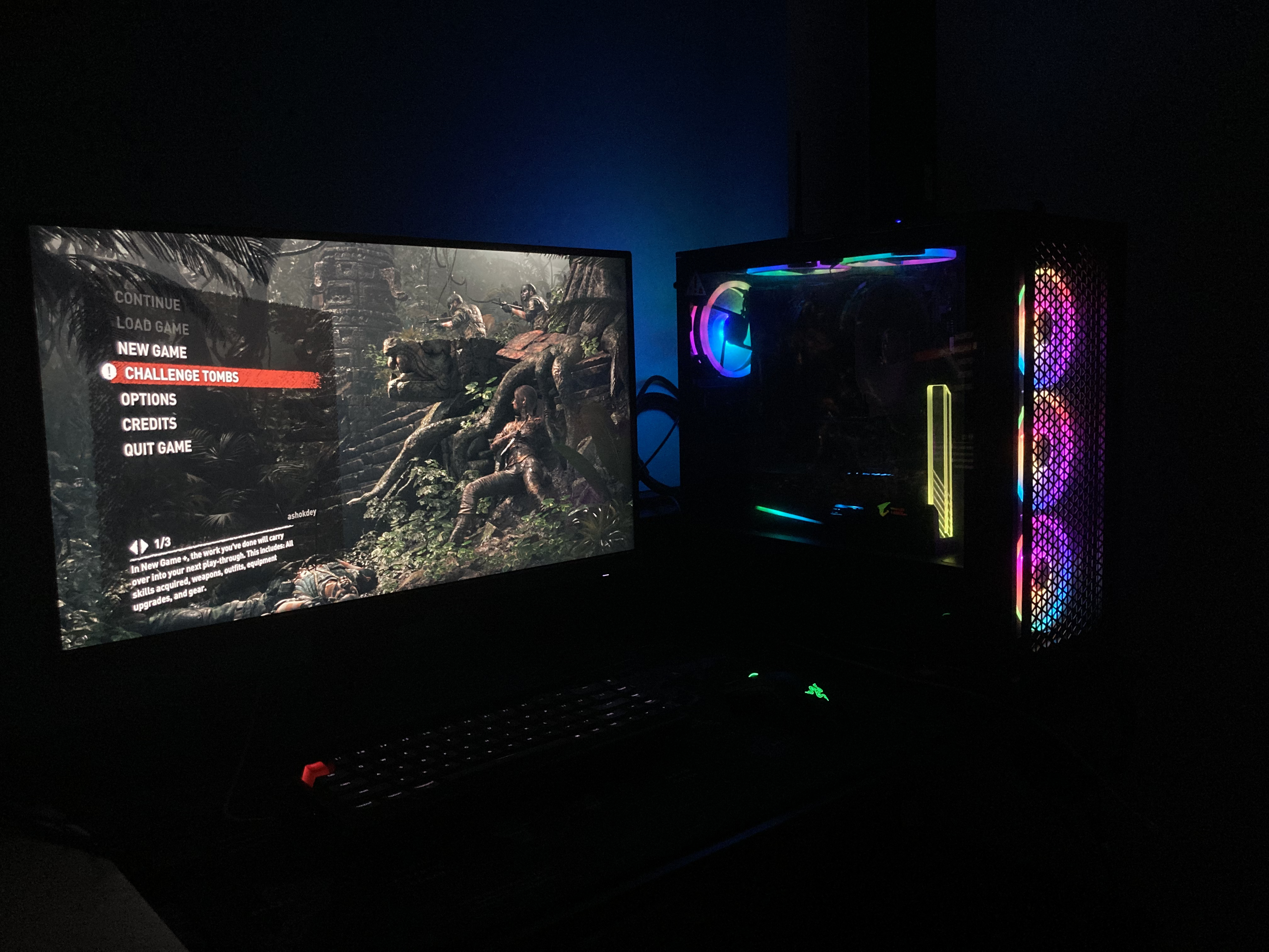 The rig running a game in a dark room, RGB fans lit, beside a monitor showing a game menu