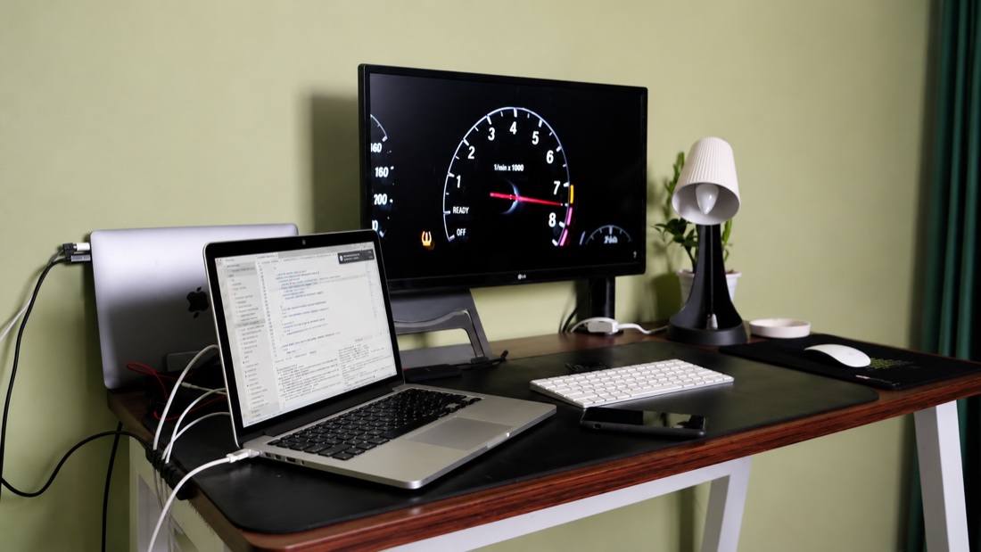 An earlier desk: an LG monitor, an open MacBook Pro showing code, a Magic Keyboard and Magic Mouse, a lamp and a plant