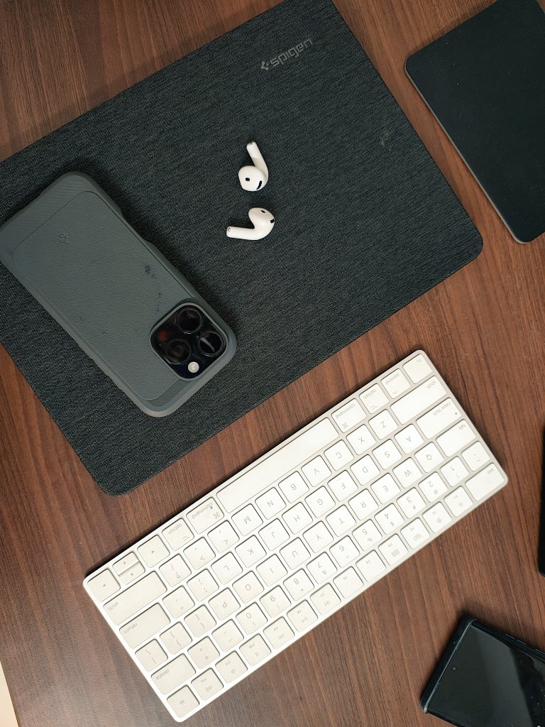 A phone in a fabric case, a pair of AirPods and an Apple Magic Keyboard on a wooden desk