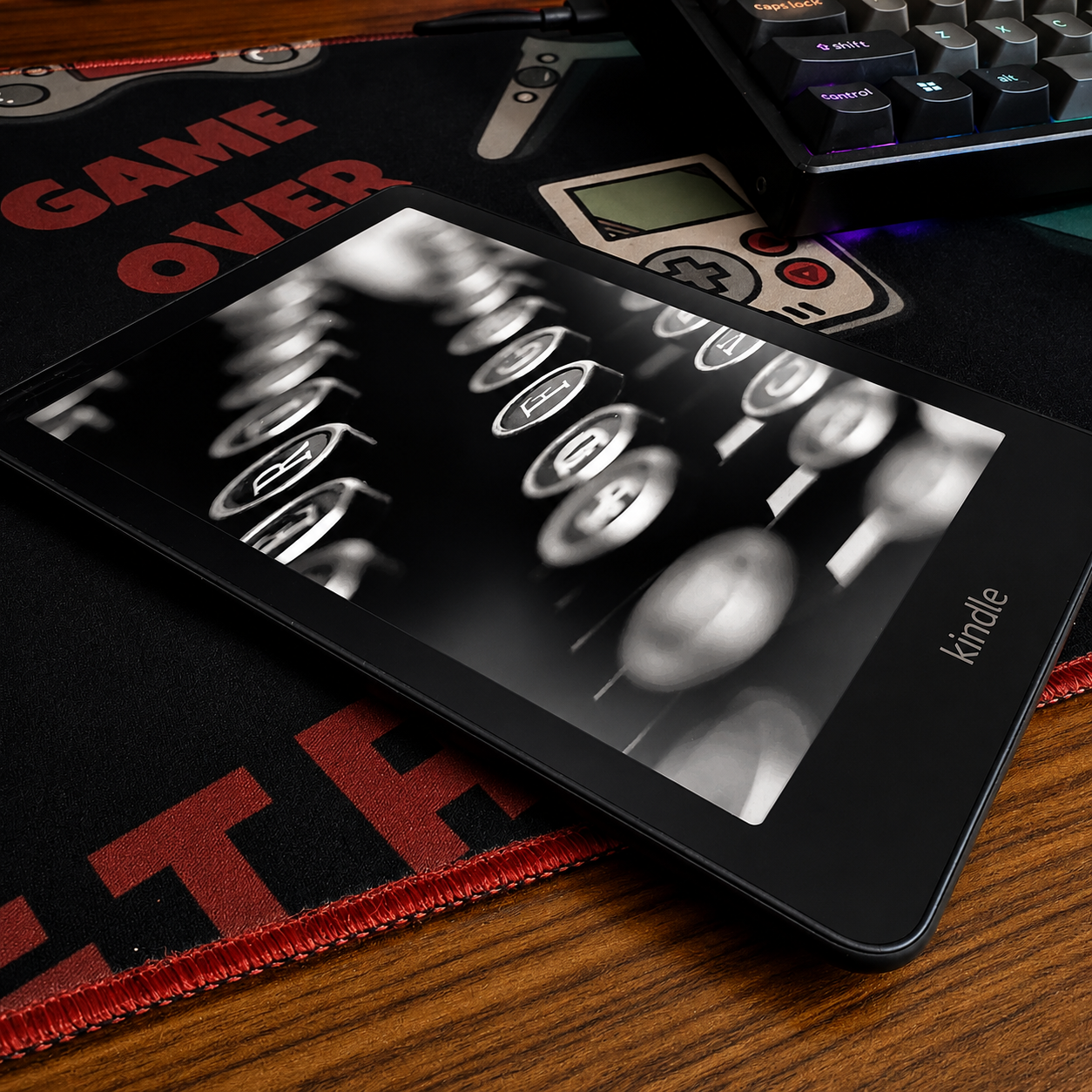 A Kindle Paperwhite showing a black-and-white typewriter cover image, resting on a Game Over desk mat beside a mechanical keyboard