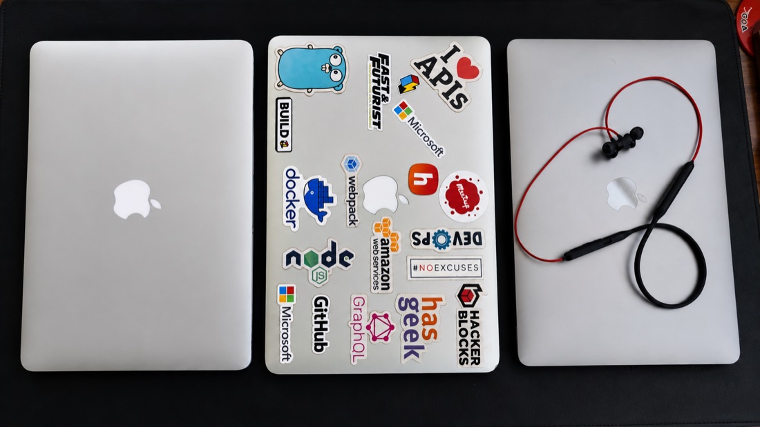 Three MacBooks side by side, the middle one covered in developer stickers, with neckband earphones resting on the third
