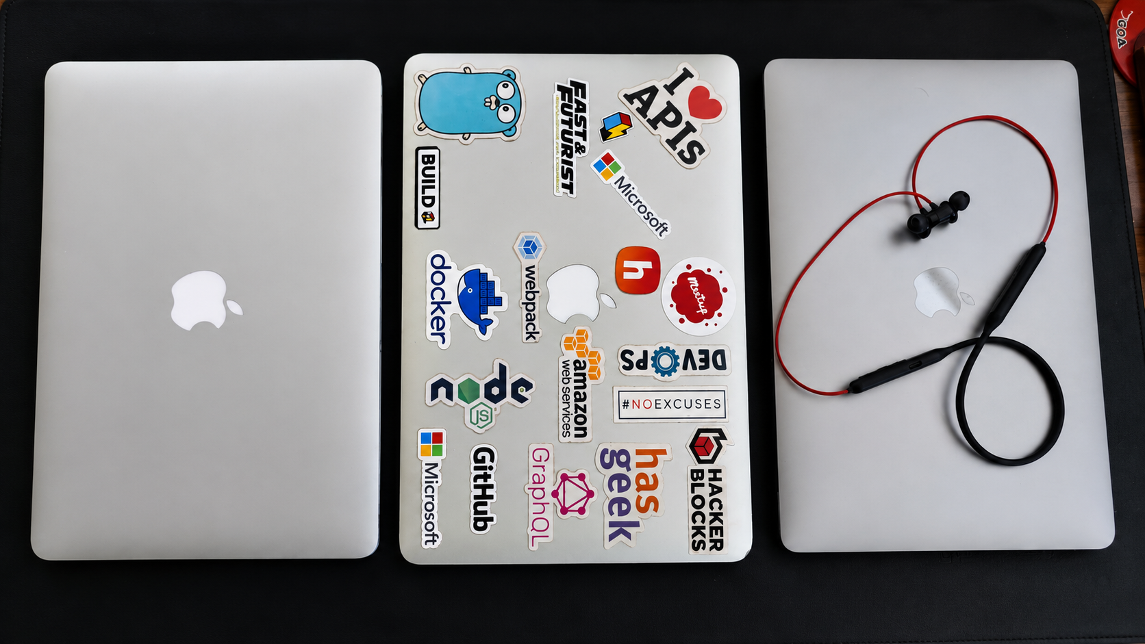 Three MacBooks side by side, the middle one covered in developer stickers, with neckband earphones resting on the third